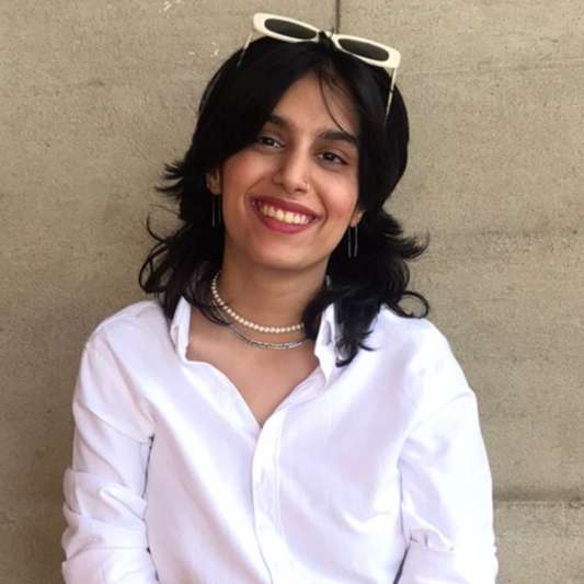 Image of Laila Dodhy, a Pakinstani woman with dark hair and a bright smile. She is wearing a white shirt, pearls, and has white sunglasses on her head.